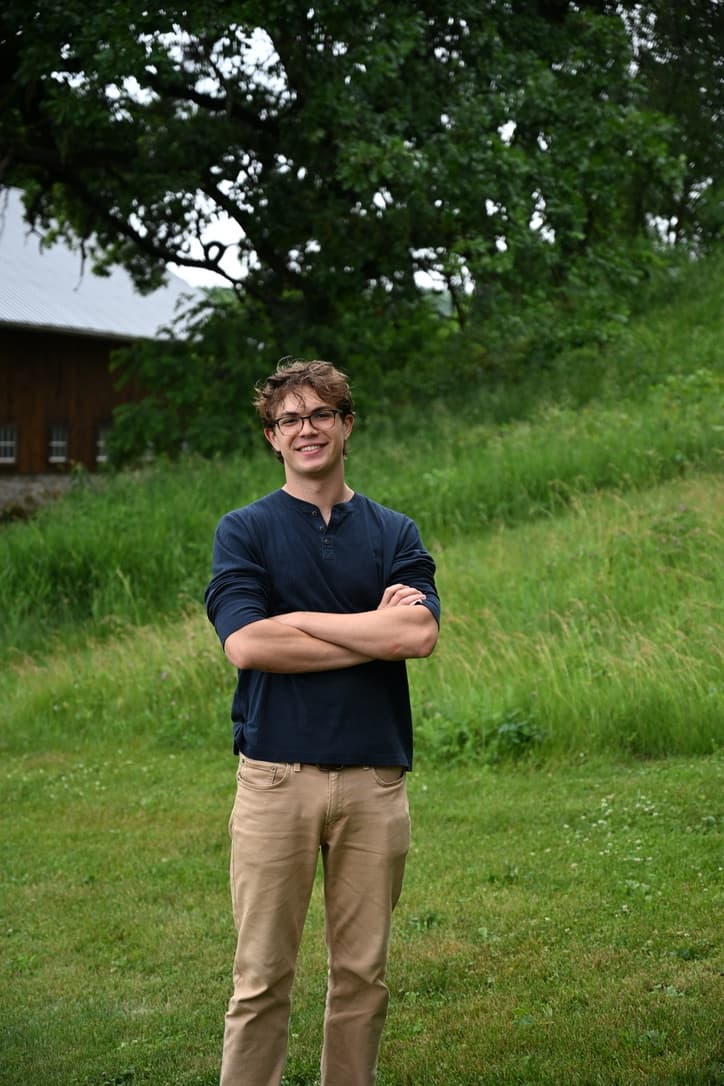 Sam, wearing glasses and a navy henley, standing with arms crossed in a field in front of a barn